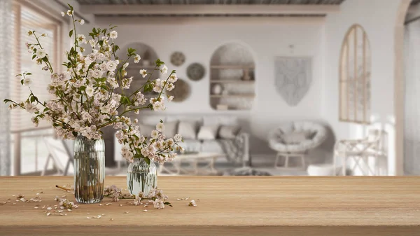Wooden table, desk or shelf close up with branches of cherry blossoms in glass vase over blurred view of bohemian living room with sofa, boho interior design concept