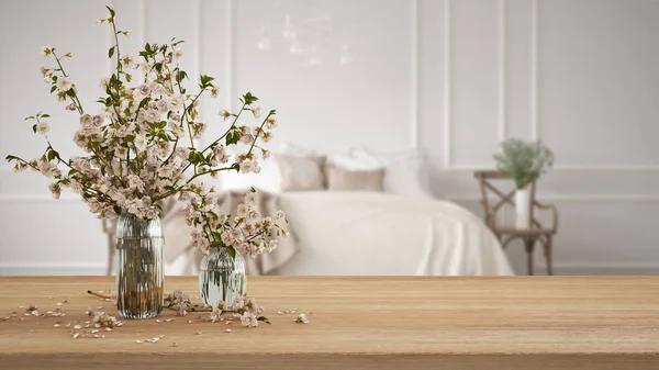 Wooden table, desk or shelf close up with branches of cherry blossoms in glass vase over blurred view of classic bedroom with soft bed, boho interior design concept