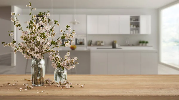Wooden table, desk or shelf close up with branches of cherry blossoms in glass vase over blurred view of modern white kitchen with island, boho interior design concept