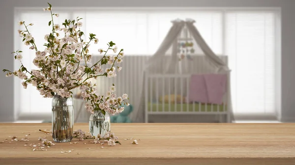 Wooden table, desk or shelf close up with branches of cherry blossoms in glass vase over blurred view of scandinavian nursery with crib, boho interior design concept