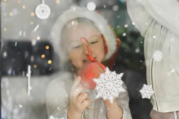 サンタの帽子をかぶった女の子が窓の雪片を見て — ストック写真