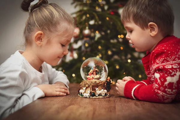 クリスマスツリーの近くにイエス キリストの誕生のシーンとガラスボールを見て子供たち — ストック写真