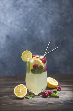 Koyu ahşap masa yüzeyinde servis edilen limonlu ve ahududulu bir bardak soğuk, serinletici içecek. Buzlu meyve ve bitki süslemeli taze kokteyller.