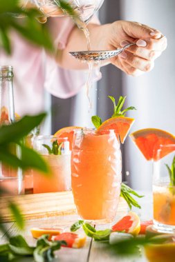 Grapefruit kokteyli hazırlayan gerçek bir kadın. İçindekiler ve açık gri masa yüzeyindeki bar aletleriyle çevrili kokteyller. İçmeye hazırım.