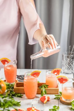 Grapefruit kokteyli hazırlayan gerçek bir kadın. İçindekiler ve açık gri masa yüzeyindeki bar aletleriyle çevrili kokteyller. İçmeye hazırım.