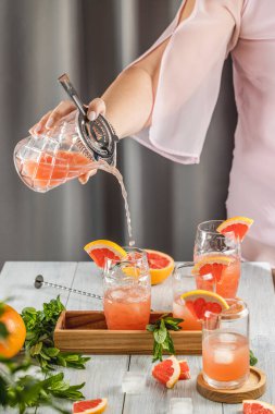 Grapefruit kokteyli hazırlayan gerçek bir kadın. İçindekiler ve açık gri masa yüzeyindeki bar aletleriyle çevrili kokteyller. İçmeye hazırım.
