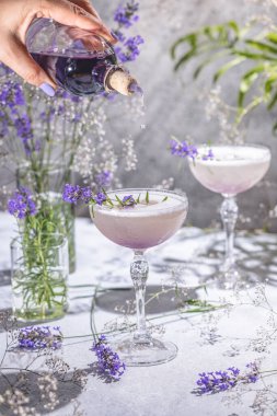 Gri masa yüzeyinde iki adet lavanta kokteyli ya da lavanta ve taze lavanta çiçekleri ile çevrili moktail. İçecekleriniz hazır..