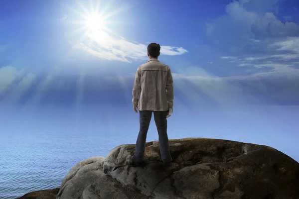 Young man standing on rock mountain and looking to the sun — Stock Photo, Image