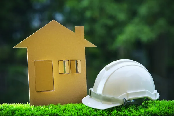 Paper home out line with safety helmet on green grass field wtih — Stock Photo, Image