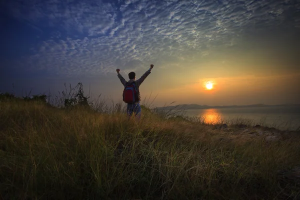 Young man standing and rising hand as victory on grass hill looking to sun above sea horizontal with dramatic colorful sky background — Stock Photo, Image