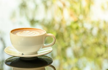Yakın plan. Cam sehpanın üzerinde Latte sanat kahve fincanı. Bulanık yaprak yansıma arkaplanı.