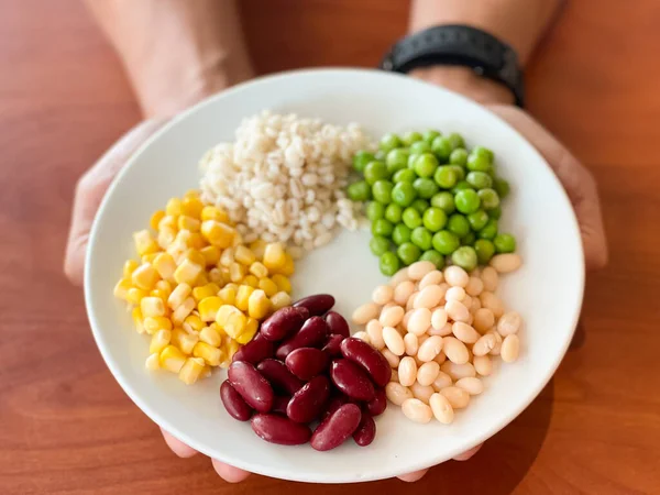 top view of navy bean and red bean and corn and  green peas and millet in white plate on hand and wood table background