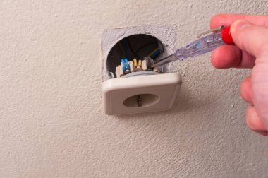 Faz bulucu tornavida yardımıyla duvardaki elektrik prizinin tamiri. Bir adamın eli faz bulucu tornavidayı tutuyor ve elektrik prizini kontrol ediyor.