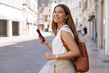 Güzel genç bir kadın akıllı telefonu tutarken ve yan bakarken şehir caddesinde yürüyor.