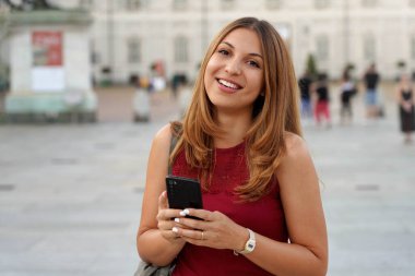 Uzun saçlı tatlı bir kadın elinde cep telefonuyla kameraya bakıyor.