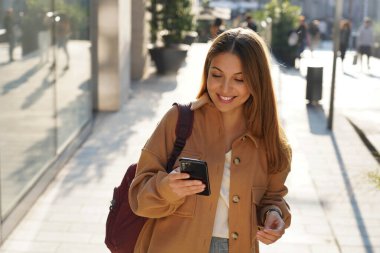 Şehirdeki alışveriş caddesinde elinde akıllı telefon tutan, gülümseyen moda kadını.