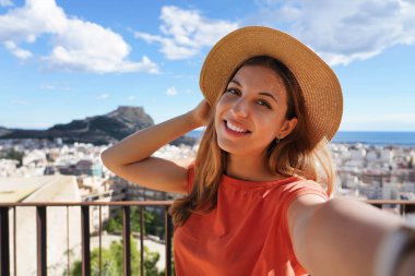 Tourist girl on Castillo de San Fernando taking selfie photo with Alicante cityscape and Mount Benacantil with Castillo de Santa Barbara on the background. Girl takes self portrait in Alicante, Spain. clipart