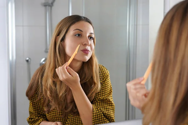 Facial hair removal. Beautiful young woman shaving her face by razor at home. Pretty woman using shaver on bathroom.