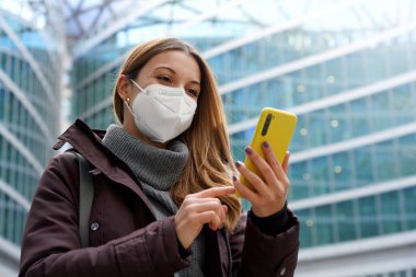 Modern şehir geçmişine sahip telefon kullanarak tıbbi maske takan bir kadının portresi. Düşük açı.