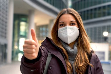 Koruyucu maske takan güzel, kendine güvenen bir kadın olan KN95 FFP2 'yi kapatın. Arka planda modern şehirle baş parmağını gösteriyor ve kameraya bakıyor.