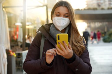 Gün batımında akıllı telefon kullanarak şehirde yürüyen tıbbi maskeli genç bir kadının portresi.