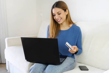 Brezilyalı güzel kız evde kanepede otururken kredi kartı ve laptopuyla online alışveriş yapıyor.