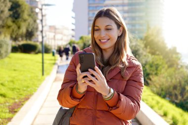 Açık havada akıllı telefon kullanan, kışlık ceket giyen ve video izleyen genç bir kadın.
