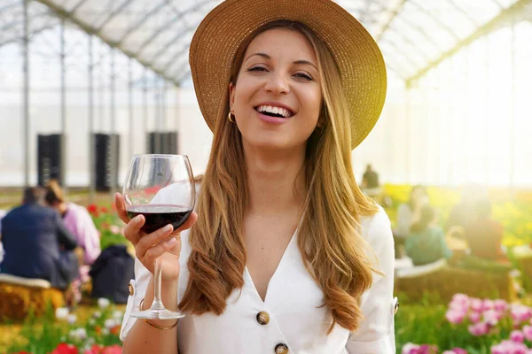 Woman looking at camera and holding glass of wine with people among flowers field drinking and chatting on the background