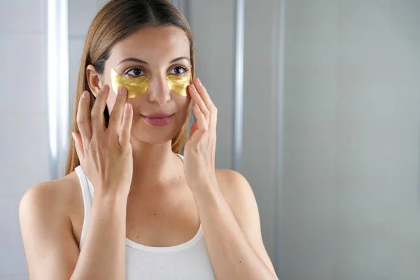 Beauty woman applying golden anti-aging under-eye mask looking herself in the mirror in the bathroom. Skin care girl touch patches of fabric mask under eyes to reduce eye bags.