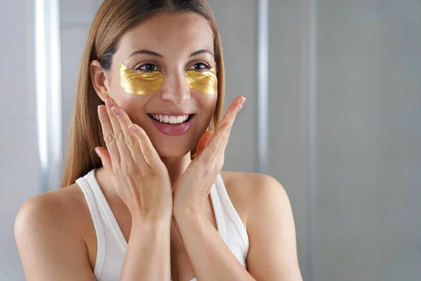 Close-up of beauty woman applying golden anti-aging under-eye mask looking herself in the mirror in the bathroom. Skin care girl touch patches of fabric mask under eyes to reduce eye bags.