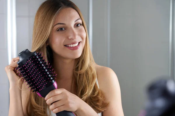 Pleased girl holds round brush hair dryer to style hair in her bathroom at home. Young woman using salon one-step hair dryer and volumizer.