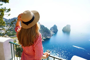 Beautiful young fashion woman with striped dress and hat in Capri Island with Faraglioni sea stack and blue crystalline water on the background, Capri, Italy
