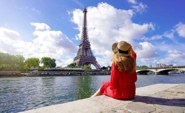 Kırmızı elbiseli ve şapkalı genç bir gezgin Seine Nehri rıhtımında oturmuş Eyfel Kulesi 'ne bakıyor. Paris' in ünlü şehir simgesi ve seyahat yeri..