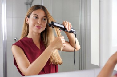 Hispanic young woman using steam straightener to style hair in the mirror on bathroom