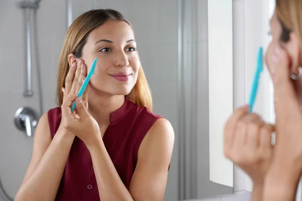 Facial hair removal. Brazilian girl shaving her face by razor at home. Beautiful young woman using razor on bathroom.