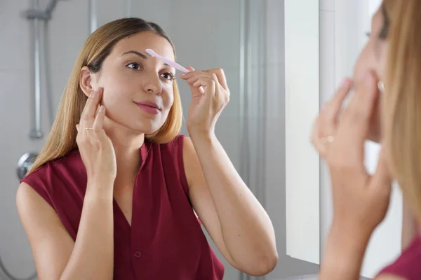 Facial hair removal. Beautiful young woman shaving her face by razor at home. Pretty woman using razor on bathroom.