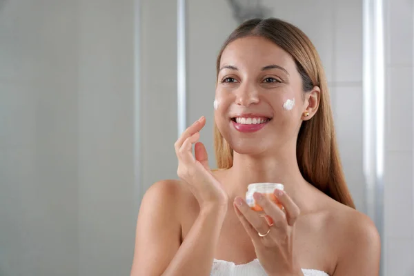 Young woman applying cream on her cheeks. Wellness treatments for teenage sensitive skin concept.