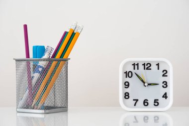 Kalemlerle dolu bir sepet, sol tarafta bir kalem ve bir düzeltici, sağ tarafında siyah sayılar ve oklar olan beyaz bir saat, ayna görüntüsü olan parlak beyaz bir masa.,