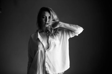 Saçları dalgalı ve kolunda gömlek olan bir kız kameraya bakar, eli yüzüne yakındır.