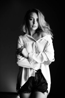 Kız, kollarını göğsünde çapraz yapıp gelişigüzel giyinmiş ve gri arka plandaki kameraya bakıyor.