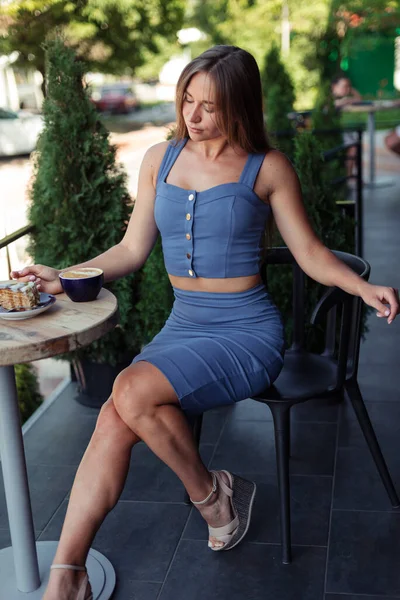 Kız terasta mavi bir üst ve kalem bir etekle oturuyor ve ayağı önde pastaya bakıyor. Yeşil Kafe.