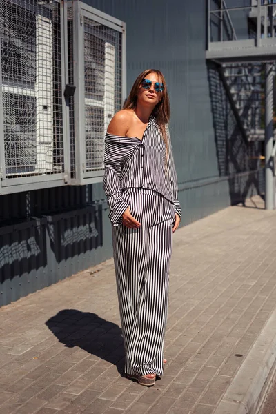 Gözlüklü genç bir kız demir bir binanın arka planında fayanslar boyunca yürüyor..