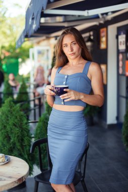 Ciddi bir kız, servilerin arasında bir kafede elinde bir bardak tutuyor..