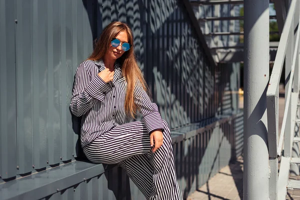 Kafkasyalı kız güneş altında gözlüklü ve geniş manşetli çizgili bir takım elbiseyle oturuyor..