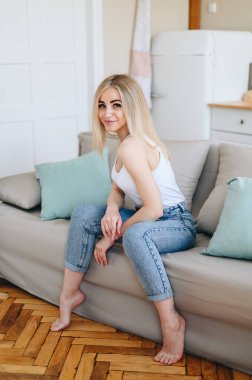 Sarışın kanepede oturur ve kameraya tatlı tatlı gülümser. Mutfakta doğal parke. Mutfakta rahatlık.