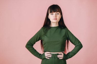 Brunette with freckles and a green suit on a pink background. The girl keeps both hands on her waist. Look seriously into the camera.