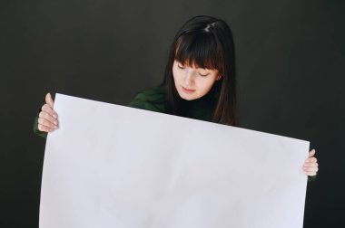 Siyah arka planda genç bir kız büyük beyaz bir sayfa tutuyor ve ona bakıyor. Bir şeyin reklamını yapma fikri.