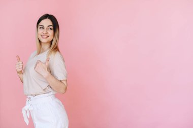 Açık renkli bir tişörtle yarım yamalak poz veren bir kız pembe arka planda iki eliyle baş parmak gösteriyor ve yan tarafa bakıyor.