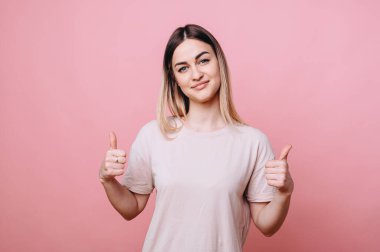 Sıradan giyinmiş bir kız, iki eliyle baş parmağını kaldırıyor ve izole bir arka planda nazikçe gülümsüyor..