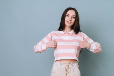 Çizgili kazaklı, neşeli, yaramaz genç bir kız, yazıtsız bir duvarda baş parmağını gösteriyor..